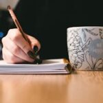 Person writing in a notebook with a floral ceramic mug on a wooden desk.